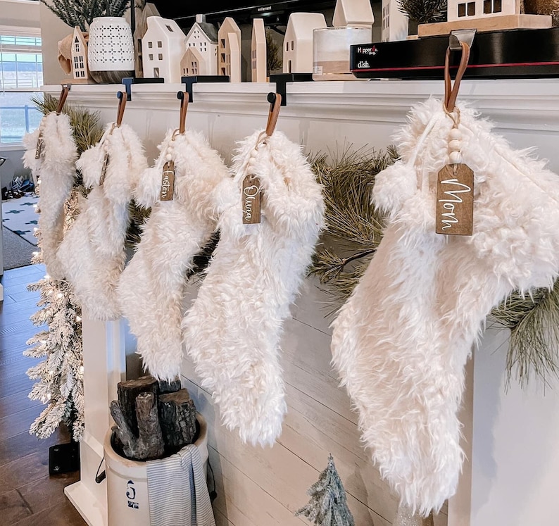 May include: Five white fuzzy Christmas stockings with wooden tags that have names written on them. The names are Olivia, Logan, Noah, and two others that are not visible. The stockings are hanging on a white mantel with a wooden background.