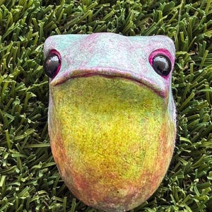 May include: A whimsical frog sculpture with a vibrant color palette. The frog's head features a mix of teal, pink, and yellow hues, with large black eyes and a wide, expressive mouth. The sculpture is set against a backdrop of green artificial grass.