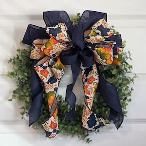 May include: A decorative bow with navy blue ribbon and a fall-themed patterned ribbon featuring orange, yellow, and green leaves. The bow is attached to a green wreath, creating a festive autumn decoration.