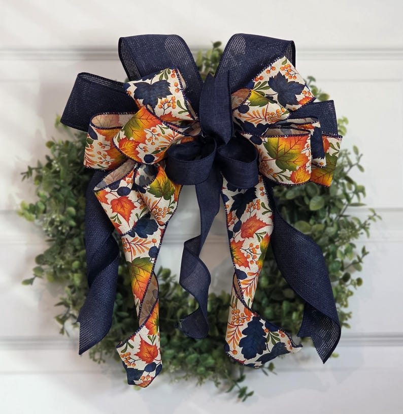 May include: A decorative bow with navy blue ribbon and a fall-themed patterned ribbon featuring orange, yellow, and green leaves. The bow is attached to a green wreath, creating a festive autumn decoration.
