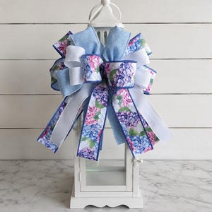 May include: A white lantern is adorned with a large bow featuring hydrangea floral print ribbons in shades of blue, pink, and purple. The bow also includes white and light blue ribbons, creating a layered effect. The lantern sits on a marble surface.