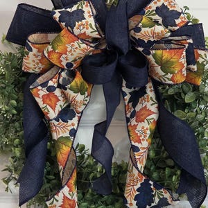 May include: A decorative wreath with a large bow. The bow is made of navy blue burlap ribbon and patterned ribbon with autumn leaves in orange, yellow, and navy blue. The wreath has a green base.