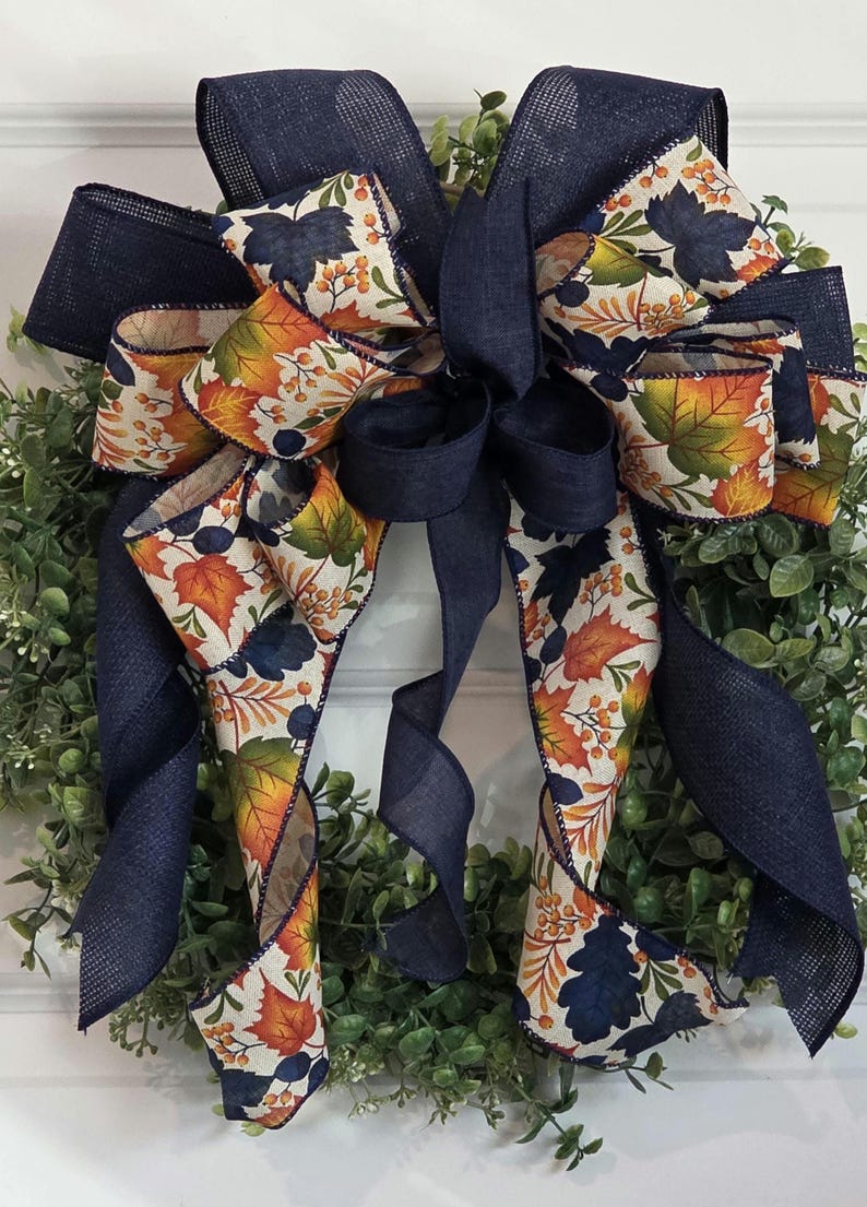 May include: A decorative wreath with a large bow. The bow is made of navy blue burlap ribbon and patterned ribbon with autumn leaves in orange, yellow, and navy blue. The wreath has a green base.