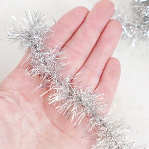 May include: A close-up of silver tinsel garland held in a hand. The tinsel is a bright, metallic silver color and has a fluffy, textured appearance. The garland is likely used for holiday decorations or crafts.