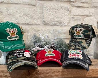 May include: Five different Christmas themed baseball caps. The caps are all green, red, black, and camouflage. The caps have different phrases on them, including "Oh Deer!" and "Merry Christmas Y'all!"