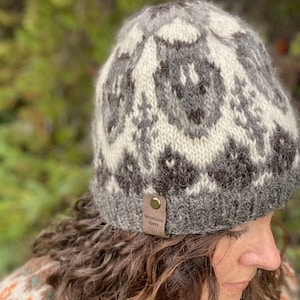 Puede incluir: Un gorro de lana tejido de color gris y blanco con un patrón geométrico. El gorro tiene una etiqueta de cuero que dice "Woodland Knits".