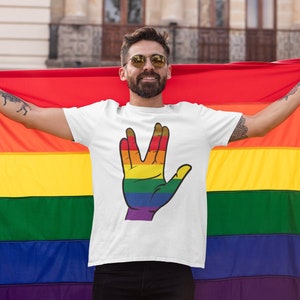 Peut inclure: Un t-shirt blanc avec une main de couleur arc-en-ciel faisant le salut Vulcain. La personne portant le t-shirt tient un drapeau arc-en-ciel de la fierté.