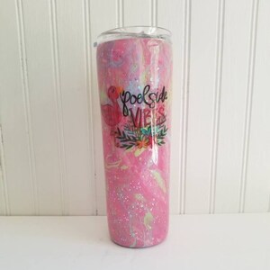 May include: A tall, insulated tumbler with a clear lid. The tumbler features a pink, white, and blue swirled design with glitter accents. The words "Poolside Vibes" are printed on the front, along with a flamingo and floral design.