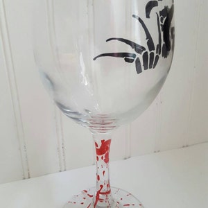 Pu&ograve; includere: Un bicchiere di vino in vetro trasparente con una silhouette nera di una mano con artigli sul lato. La base del bicchiere &egrave; dipinta con vernice rossa a spruzzo.