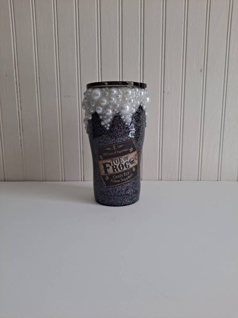 May include: A black and silver glitter tumbler with a white pearl rim and a label that reads "Toe of Frog, Crazy Elf Potion Supplier".