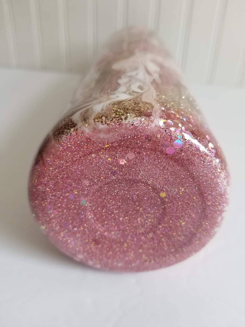 May include: A pink glitter tumbler with white and gold swirls. The tumbler is filled with pink glitter and has a clear lid.