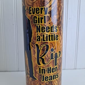 May include: A gold and brown tumbler with a black silhouette of a person and the text "Every Girl Needs a Little Rip in Her Jeans".