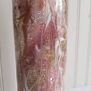 May include: A pink and gold glitter tumbler with white swirls. The tumbler has a clear, smooth surface and is filled with glitter.