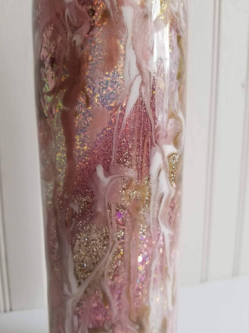 May include: A pink and gold glitter tumbler with white swirls. The tumbler has a clear, smooth surface and is filled with glitter.
