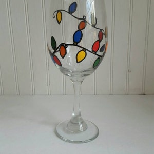 May include: A clear glass wine glass with a hand-painted design of colorful Christmas lights. The lights are strung on a black wire and are in various shades of red, blue, yellow, and green.
