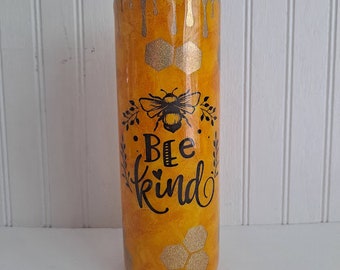 May include: A yellow and gold tumbler with a bee and honeycomb design. The tumbler has the text "Bee Kind" with a heart in the word "Bee".