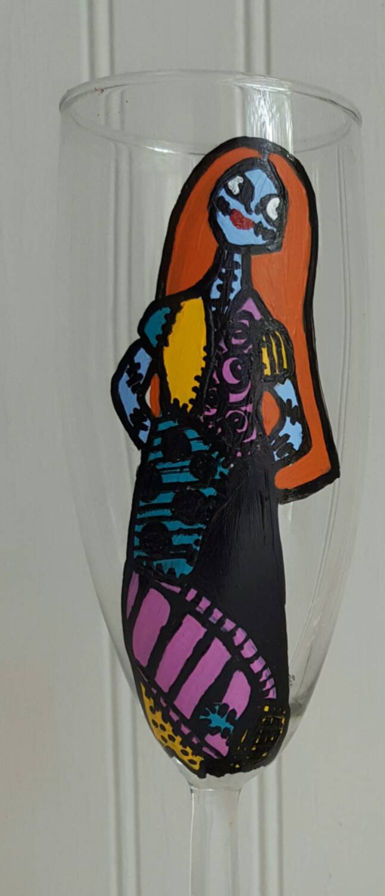 Pode incluir: Uma ta&ccedil;a de vinho pintada &agrave; m&atilde;o com uma ilustra&ccedil;&atilde;o colorida de Sally de O Estranho Mundo de Jack. A ta&ccedil;a &eacute; transparente e tem um caule.