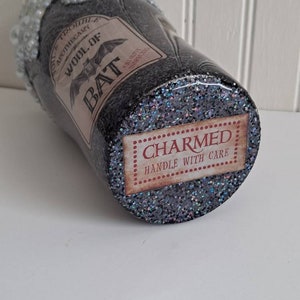 May include: A black glitter tumbler with a white foamy top and a label that reads "Old Trouble's Apothecary Wool of Bat" with a bat graphic. The bottom of the tumbler has a label that reads "CHARMED HANDLE WITH CARE".