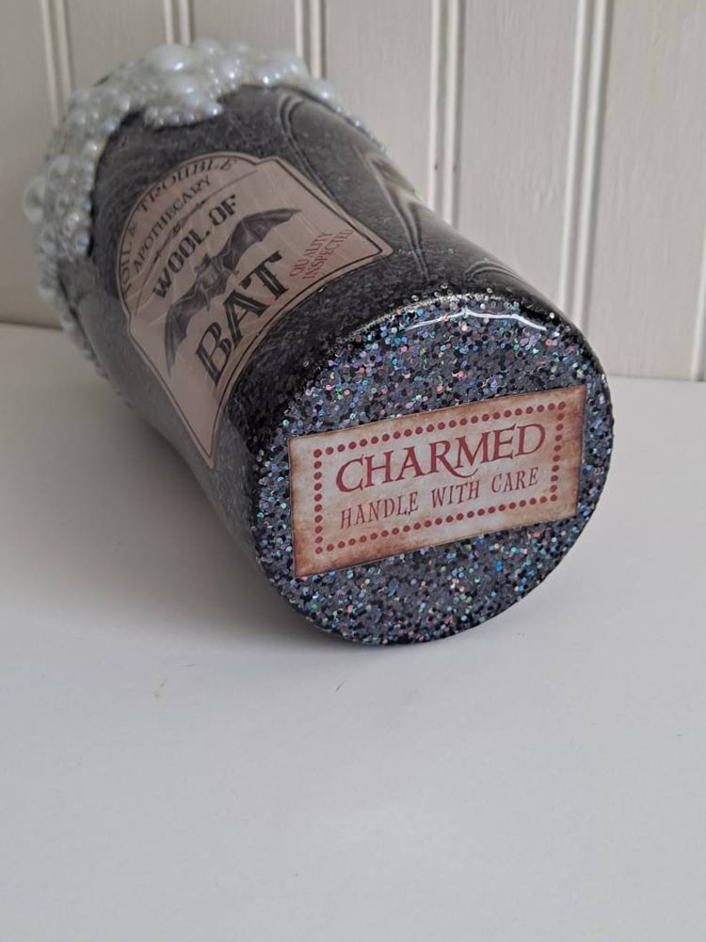 May include: A black glitter tumbler with a white foamy top and a label that reads "Old Trouble's Apothecary Wool of Bat" with a bat graphic. The bottom of the tumbler has a label that reads "CHARMED HANDLE WITH CARE".