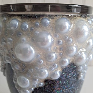 May include: A black and silver glitter tumbler with a white pearl-like design. The tumbler has a black rim and a silver glitter base.
