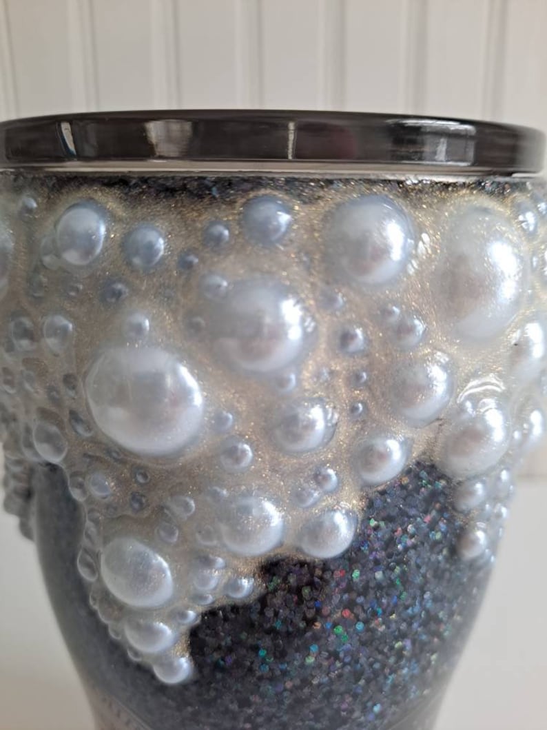 May include: A black and silver glitter tumbler with a white pearl-like design. The tumbler has a black rim and a silver glitter base.