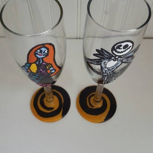 Pode incluir: Duas ta&ccedil;as de champanhe pintadas &agrave; m&atilde;o com desenhos em espiral preto e laranja na base. Uma ta&ccedil;a apresenta uma imagem de desenho animado de Sally do filme O Estranho Mundo de Jack, e a outra ta&ccedil;a apresenta uma imagem de desenho animado de Jack Skellington do filme O Estranho Mundo de Jack.