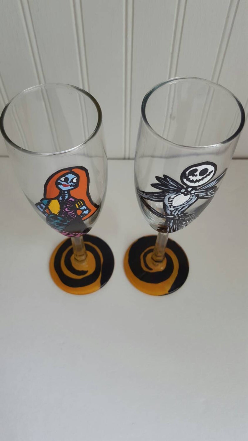 Pode incluir: Duas ta&ccedil;as de champanhe pintadas &agrave; m&atilde;o com desenhos em espiral preto e laranja na base. Uma ta&ccedil;a apresenta uma imagem de desenho animado de Sally do filme O Estranho Mundo de Jack, e a outra ta&ccedil;a apresenta uma imagem de desenho animado de Jack Skellington do filme O Estranho Mundo de Jack.