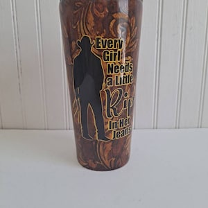 May include: A brown and gold tumbler with a silhouette of a person wearing a cowboy hat and the text "Every Girl Needs a Little  Rip In Her Jeans".