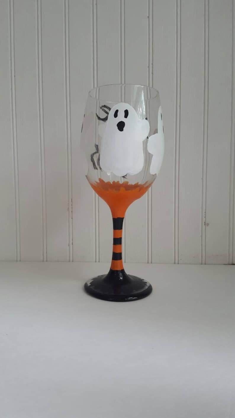 May include: A clear glass wine glass with a black stem and base. The glass is decorated with orange and black stripes and three white ghosts with black eyes.