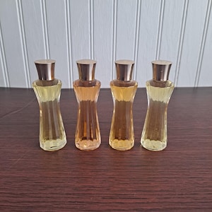 May include: Four small glass bottles with gold caps and a yellow liquid inside. The bottles are lined up in a row on a wooden surface.