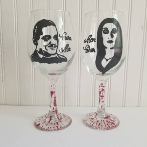 May include: Two wine glasses with black and white illustrations of a man and a woman. The glasses have red paint splatters on the stems and bases. The text "Cara Mia" is written on the glass with the man and "Mon Cher" is written on the glass with the woman.