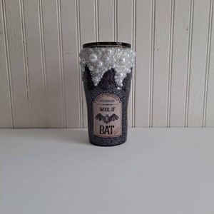 May include: A black and silver glitter tumbler with a white pearl rim. The tumbler has a label that reads "Toil & Trouble Apothecary" and "Wool of Bat" with a bat graphic.