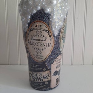 May include: A black and silver glitter tumbler with a white, pearl-like finish. The tumbler features various vintage-style labels with text including "The Apothecary Est. 1766", "Amortentia", "Toe of Frog", and "Bat".