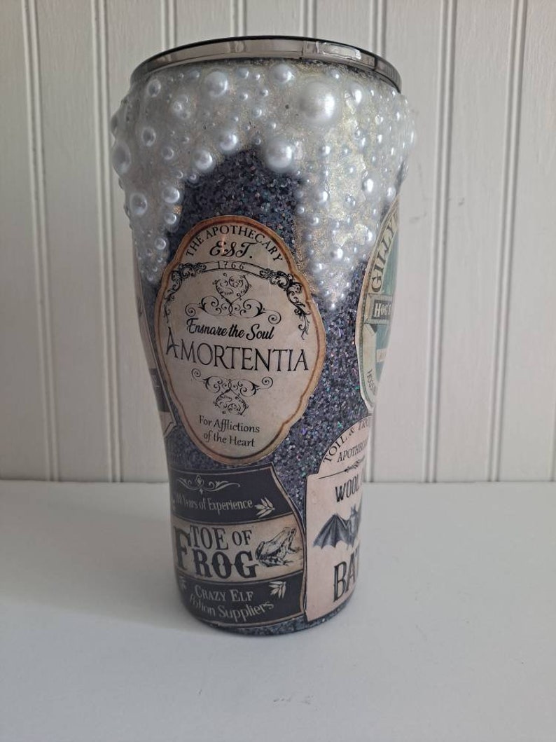 May include: A black and silver glitter tumbler with a white, pearl-like finish. The tumbler features various vintage-style labels with text including "The Apothecary Est. 1766", "Amortentia", "Toe of Frog", and "Bat".