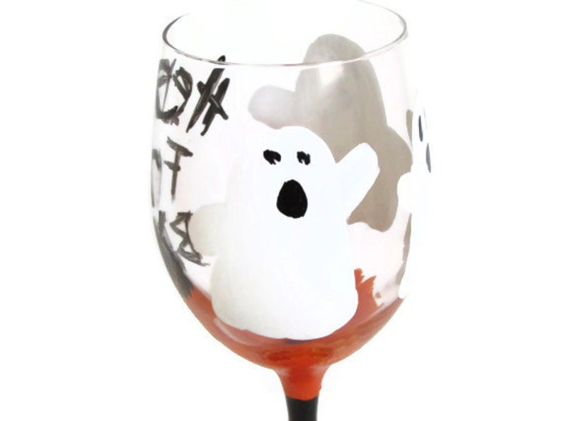 May include: A clear glass wine glass with a black and white ghost design and orange paint around the base. The glass has the words "Boo!" written on it.