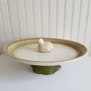 May include: A white ceramic cake stand with a green base. The stand has a decorative rim and a small ceramic bird figurine on top.