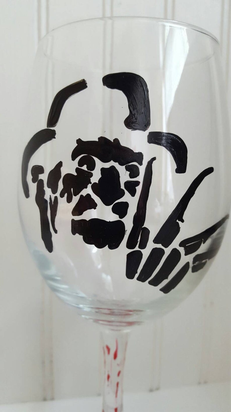 Pu&ograve; includere: Un bicchiere da vino in vetro trasparente con una silhouette nera del viso e della mano di un personaggio di un film horror. Il gambo del bicchiere &egrave; dipinto con strisce rosse.