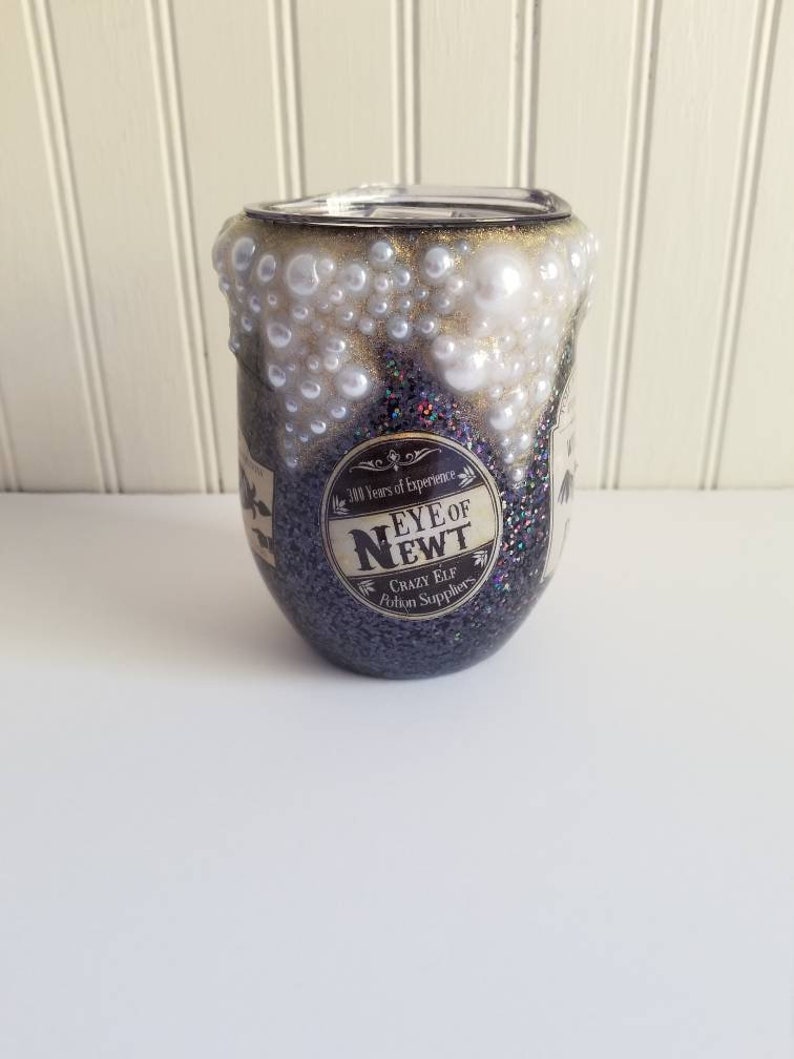 May include: A black glitter tumbler with a gold rim and white pearl accents. The tumbler has a label that reads "Eye of Newt" with the text "300 Years of Experience" and "Crazy Elf Potion Suppliers".