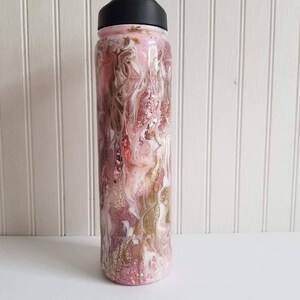May include: A pink and gold glitter water bottle with a black lid. The bottle has a swirling pattern of white, pink, and gold glitter.