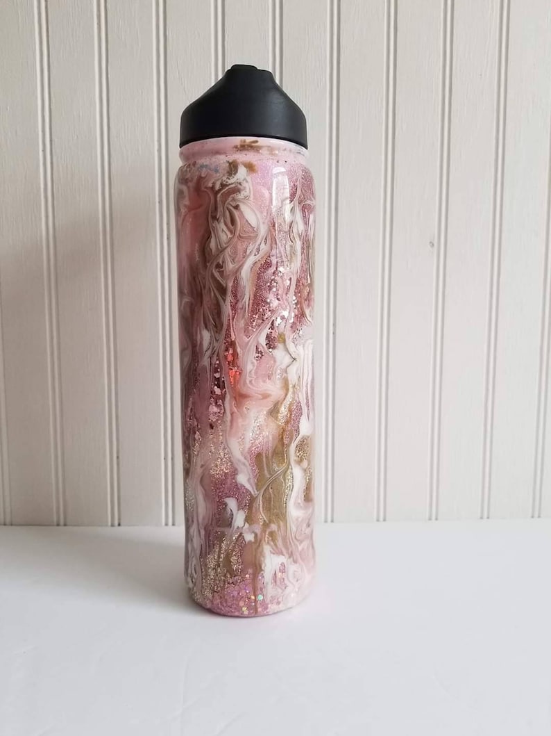 May include: A pink and gold glitter water bottle with a black lid. The bottle has a swirling pattern of white, pink, and gold glitter.