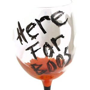 May include: A clear glass wine glass with a black painted design that says "Here For Booze" in a handwritten style. The glass is filled with a bright orange liquid.