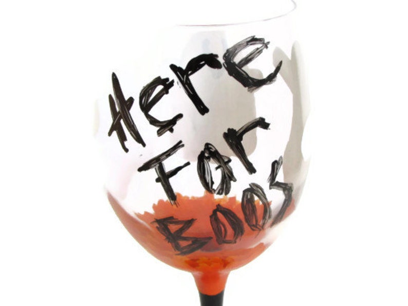 May include: A clear glass wine glass with a black painted design that says "Here For Booze" in a handwritten style. The glass is filled with a bright orange liquid.