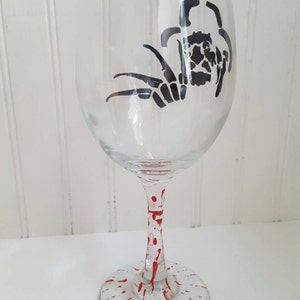 Pu&ograve; includere: Un bicchiere da vino in vetro trasparente con una silhouette nera di un ragno sul lato. Il gambo del bicchiere &egrave; decorato con schizzi di vernice rossa.