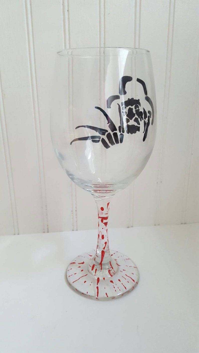 Pu&ograve; includere: Un bicchiere da vino in vetro trasparente con una silhouette nera di un ragno sul lato. Il gambo del bicchiere &egrave; decorato con schizzi di vernice rossa.