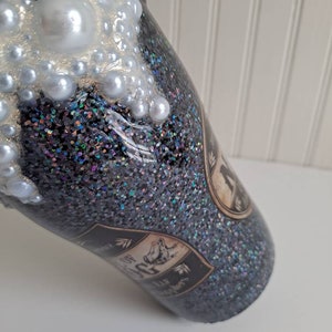 May include: A black glitter tumbler with a white pearl-like design on the top. The tumbler has a black and white label with the text "The House of the Rising Sun" printed on it.