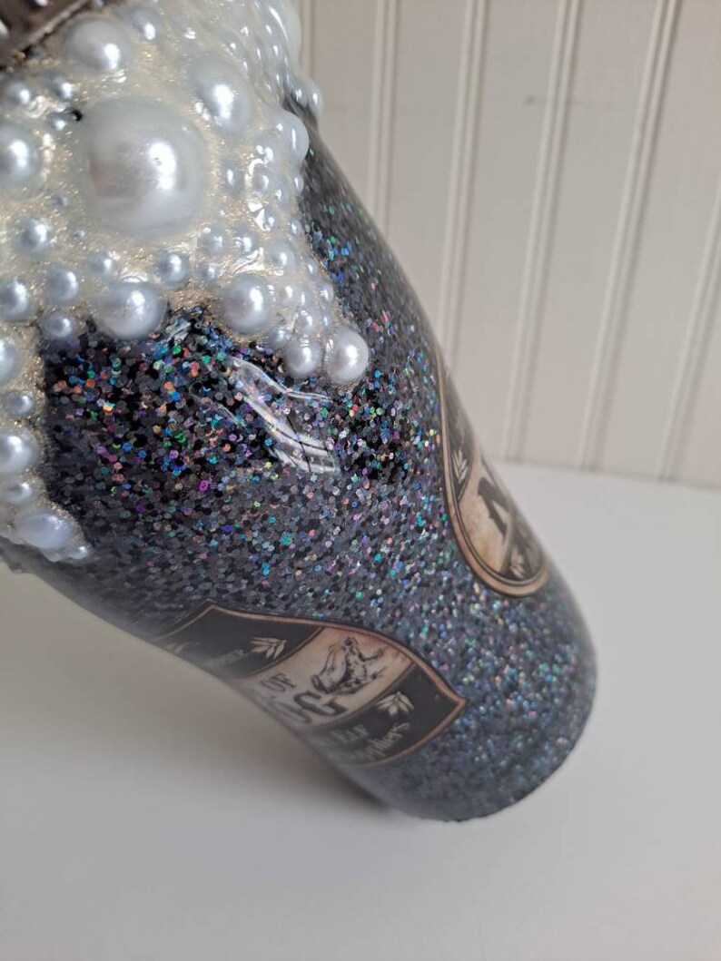 May include: A black glitter tumbler with a white pearl-like design on the top. The tumbler has a black and white label with the text "The House of the Rising Sun" printed on it.