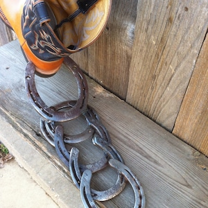 May include: A brown leather cowboy boot with a black leather pull tab hangs upside down over a stack of five silver horseshoes.