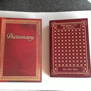 May include: Two hardcover books with red covers. The larger book is labeled "Dictionary" in gold script. The smaller book is titled "Probability & Truth" and lists "Ms. May Bee" as the author. Both books have gold decorative borders.