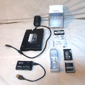May include: An assortment of electronic devices and accessories, including a black Apava card reader, a RadioShack battery charger, and various batteries. Cables and connectors are also visible. The items are arranged on a light-colored surface.