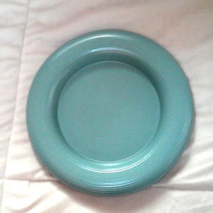 May include: A round, teal-colored plate with a slightly raised rim and a textured, ridged design. The plate has a smooth, flat center and a wide, textured border. The plate is made of a solid, opaque material.